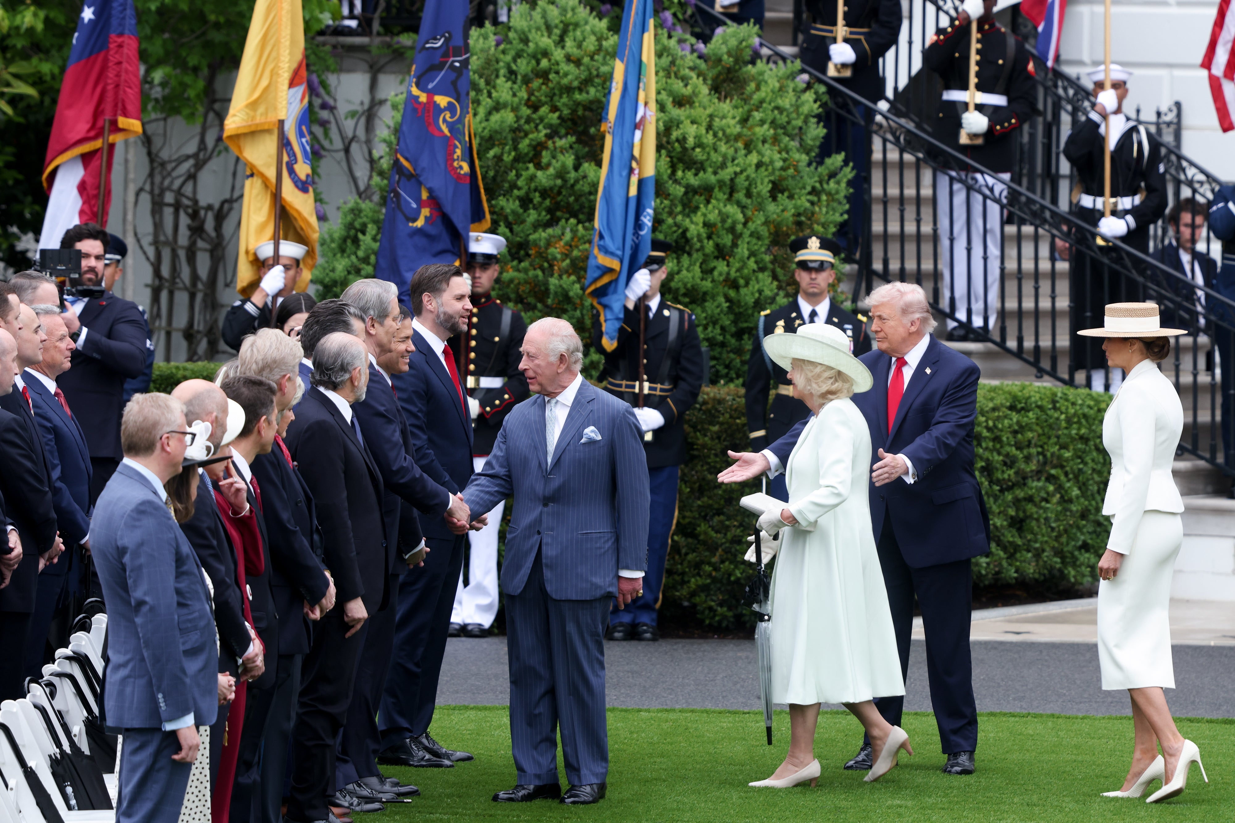 Gruppe av dignitærer på et utendørsarrangement, inkludert en mann i formell dress og en kvinne i en elegant kjole med en hatt, hilser deltakere