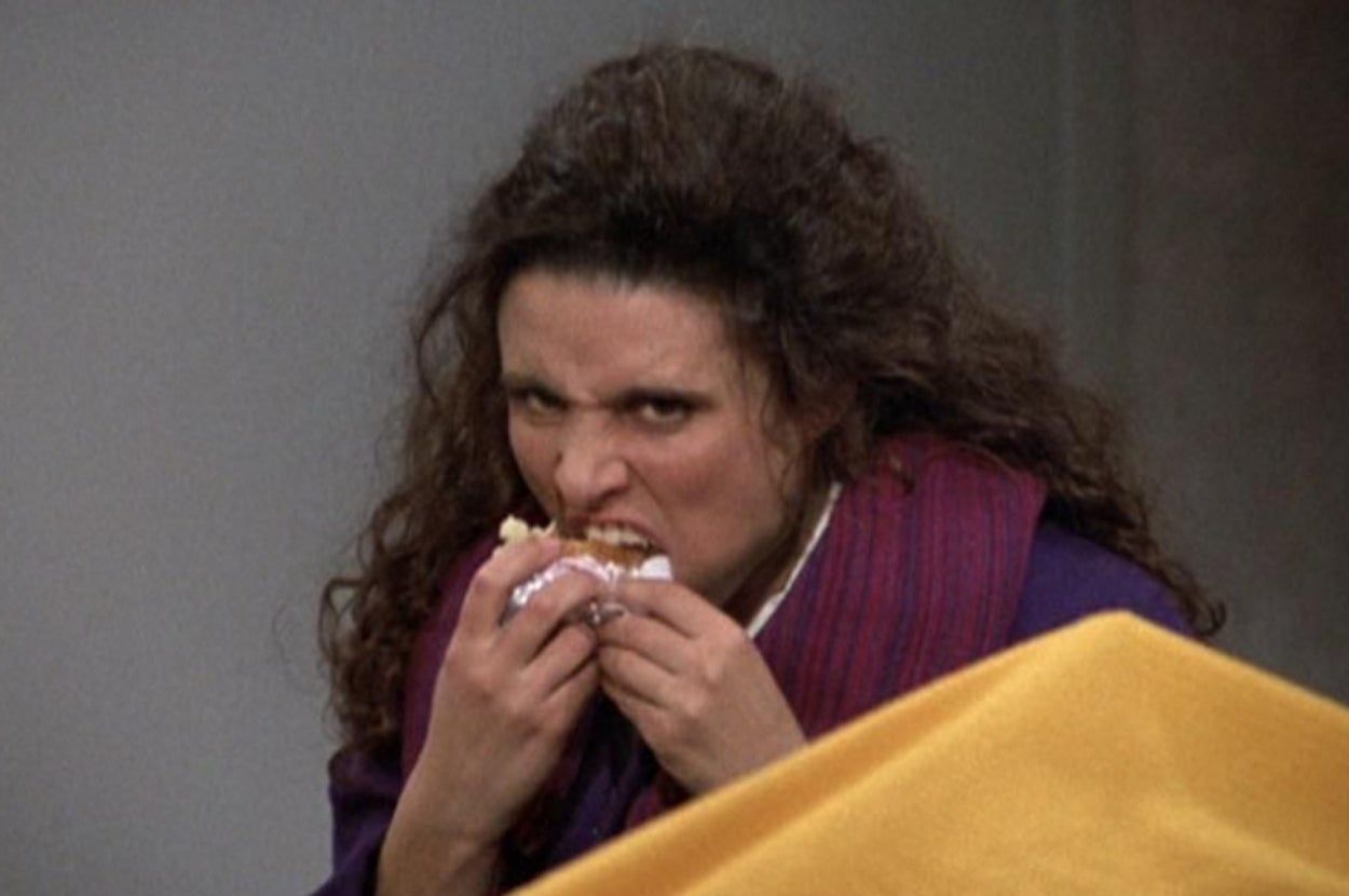 A person with curly hair eagerly biting into a sandwich, showcasing an intense and humorous expression