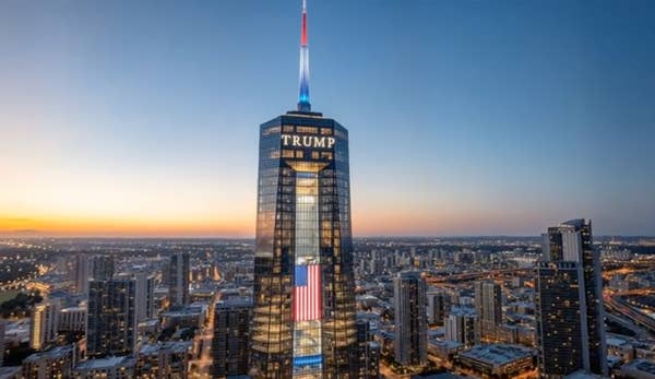 A tall skyscraper with "Trump" at the top stands prominently among a cityscape, with a spire lit in blue, white, and red