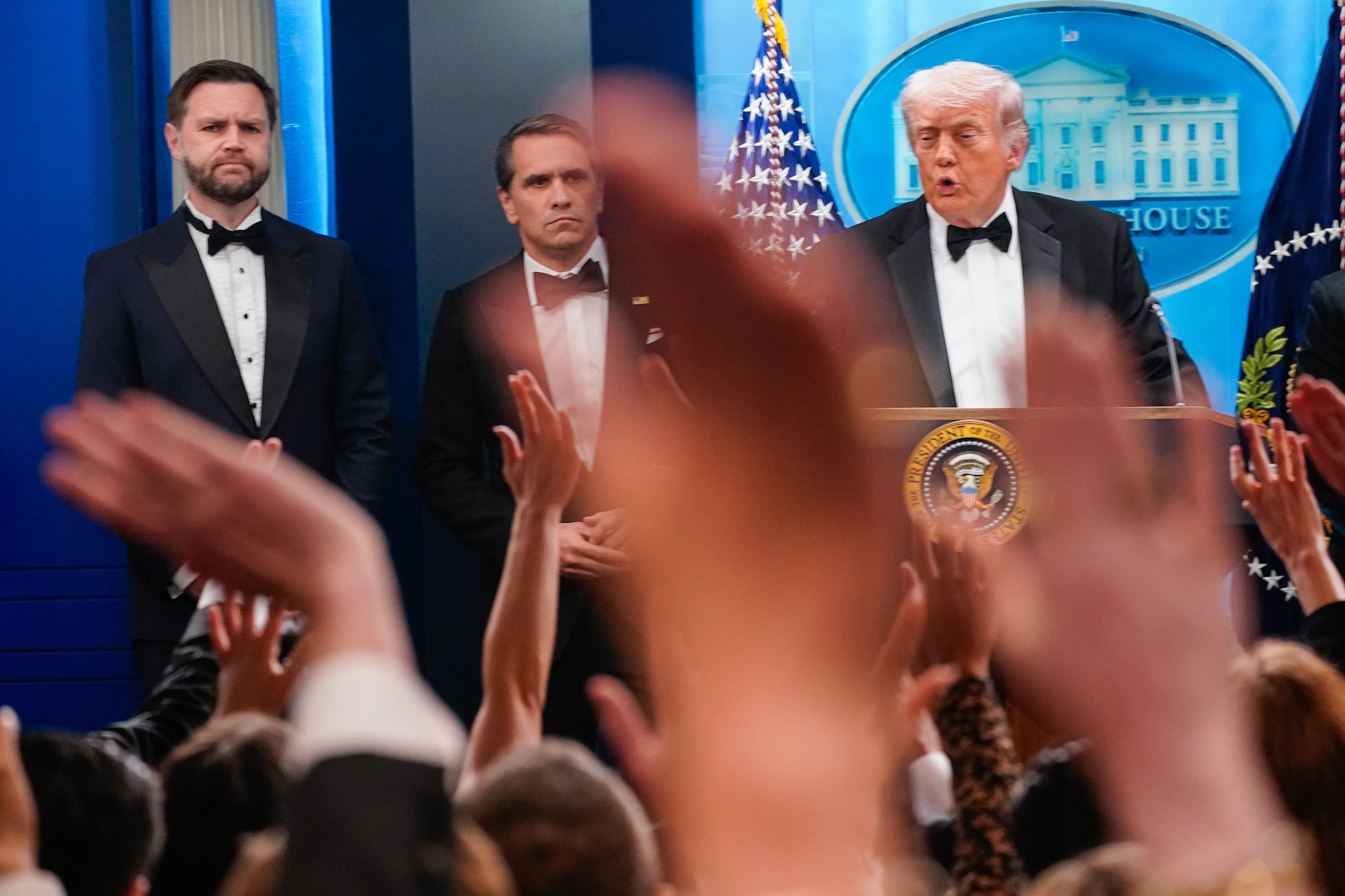 En mann i smoking taler ved et podium med en presidentsegl, to andre menn i smoking står ved siden av ham, med hendene hevet fra et publikum foran