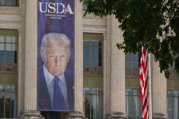 A large banner of a person is displayed on a USDA building, featuring their serious expression. An American flag is visible beside it