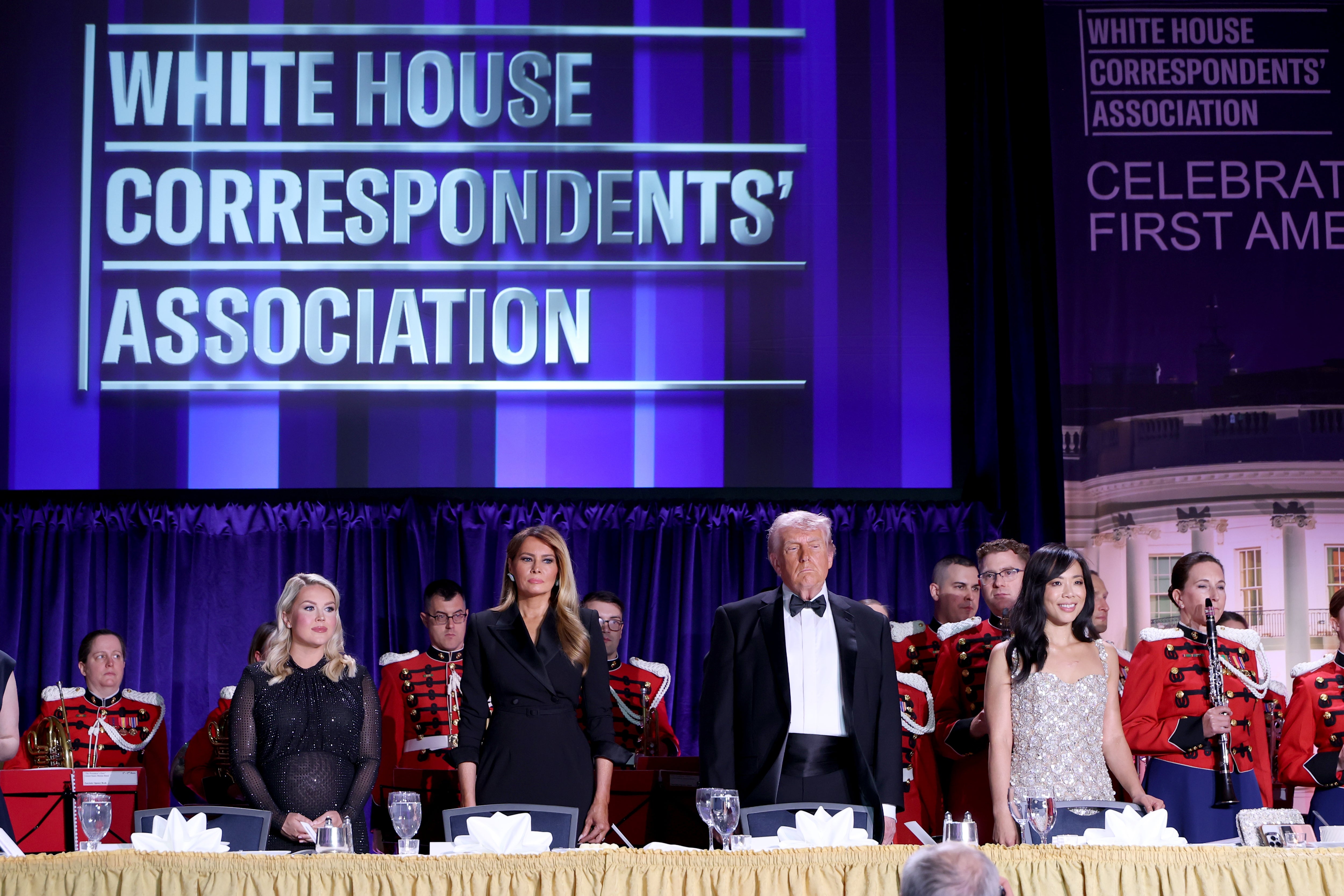 Folk på en formell begivenhet med en "White House Correspondents' Forening" skilt. De står foran musikere iført røde uniformer