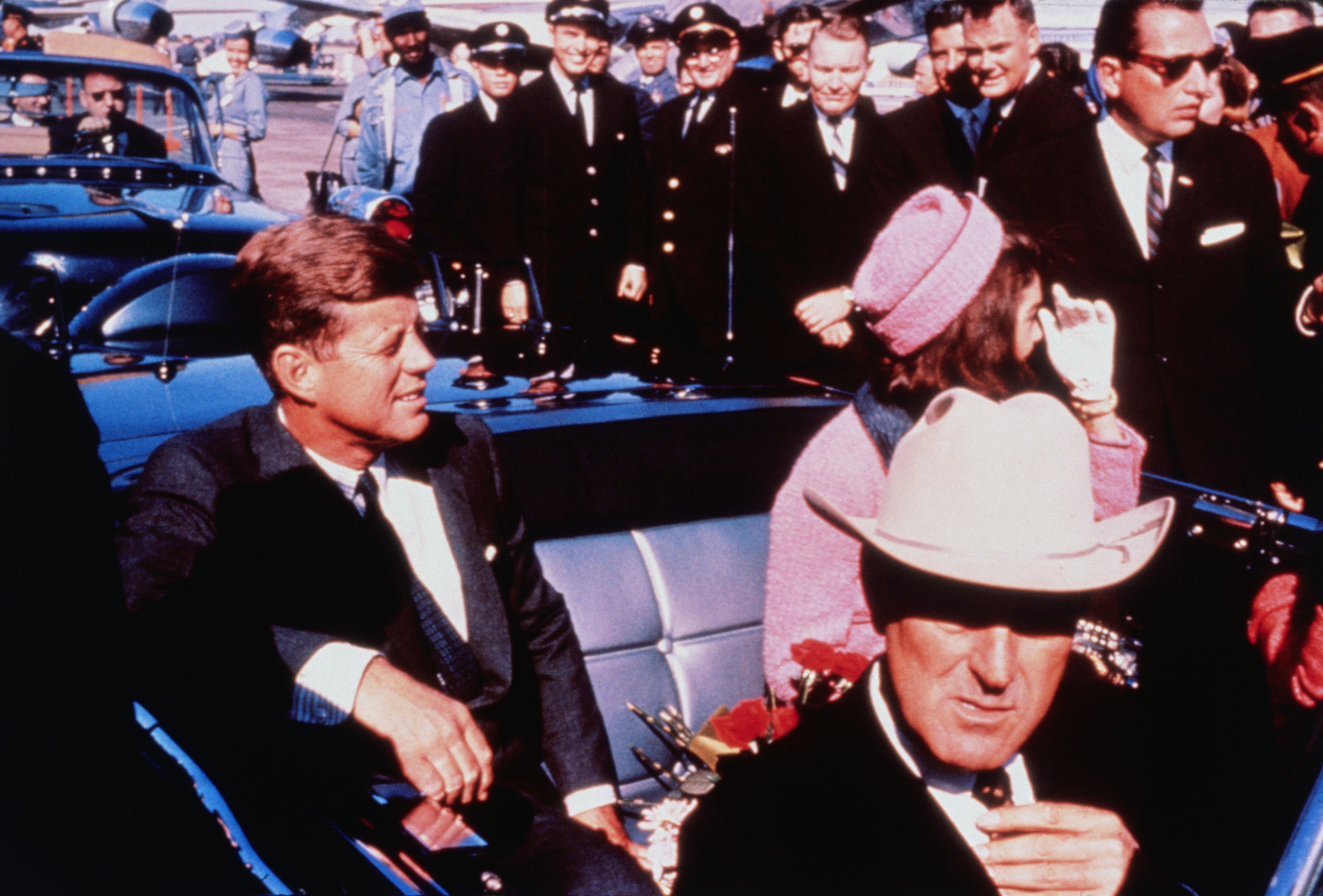Et vintagebilde av John F. Kennedy i en cabriolet sammen med andre, inkludert en kvinne i en stilig hatt og en mann i en cowboyhatt, omgitt av tjenestemenn