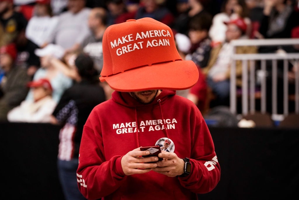 Person wearing oversized "Make America Great Again" chapeau  and hoodie, looking astatine  their telephone  successful  a crowded setting