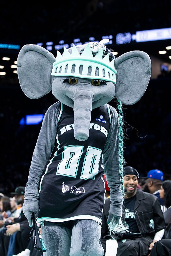 A sports mascot dressed arsenic  a stylized elephant with a hoops  jersey and a crown-like headpiece astatine  an indoor sports event