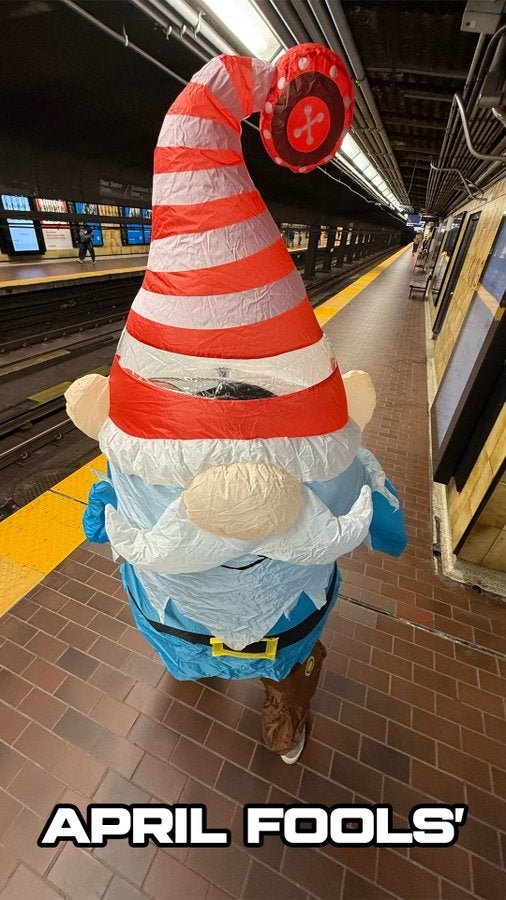 Person wearing an inflatable gnome costume with a gangly  reddish  and achromatic  chapeau  stands connected  a subway platform. Text reads "April Fools'"
