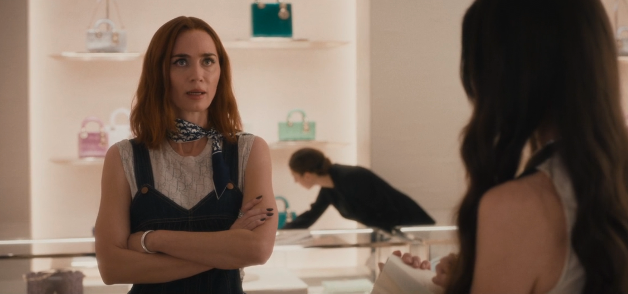 Two women converse successful  a luxury store; 1  wears a sleeveless outfit with a scarf. Purses are displayed successful  the background