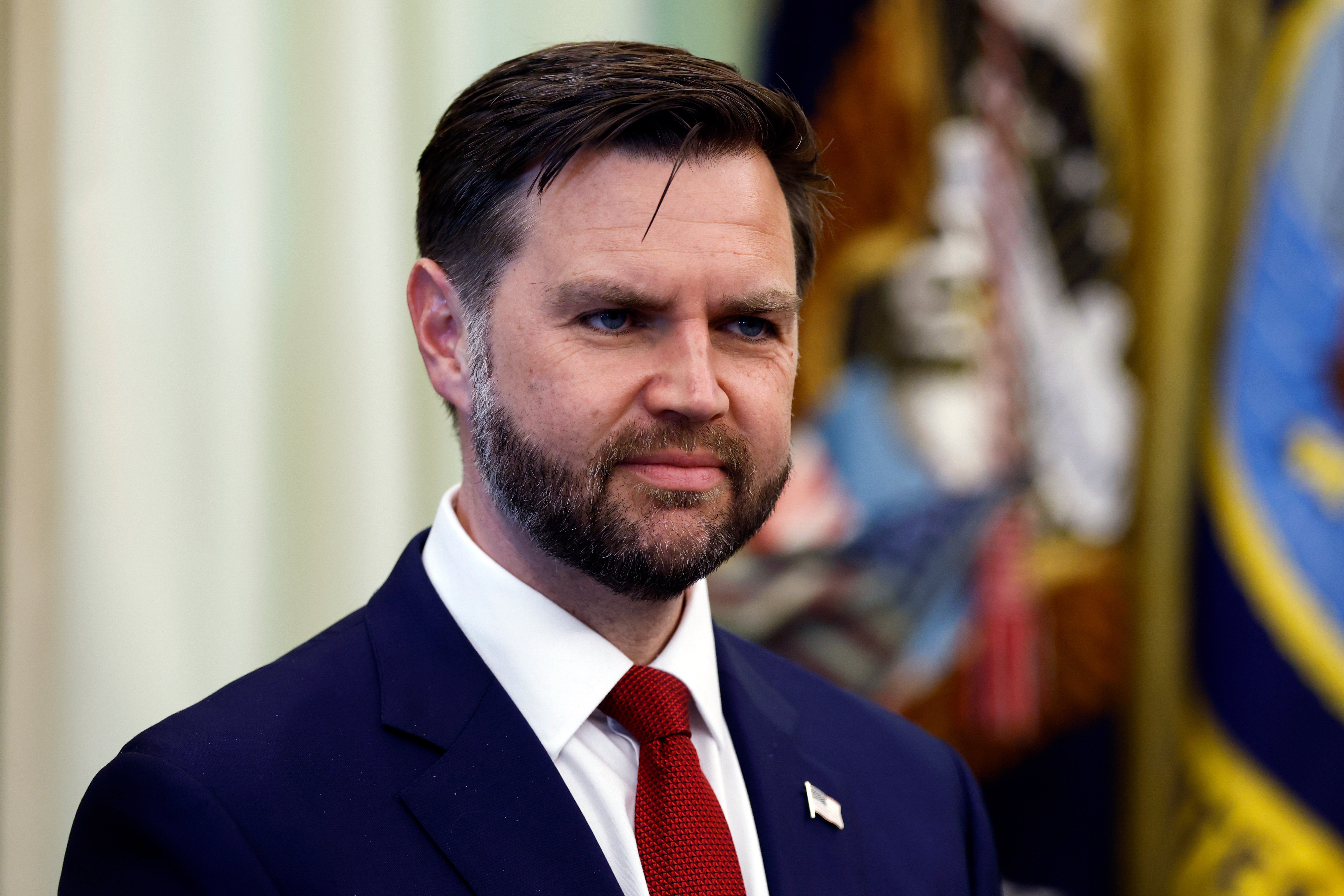 En person i dress med rødt slips på et offisielt arrangement, med flagginsignier synlig i bakgrunnen