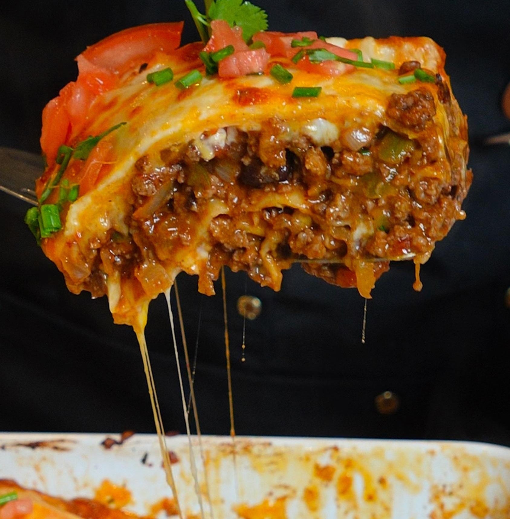 A close-up of a scoop of cheesy, baked lasagna with meat sauce, topped with diced tomatoes and herbs, being lifted from a dish