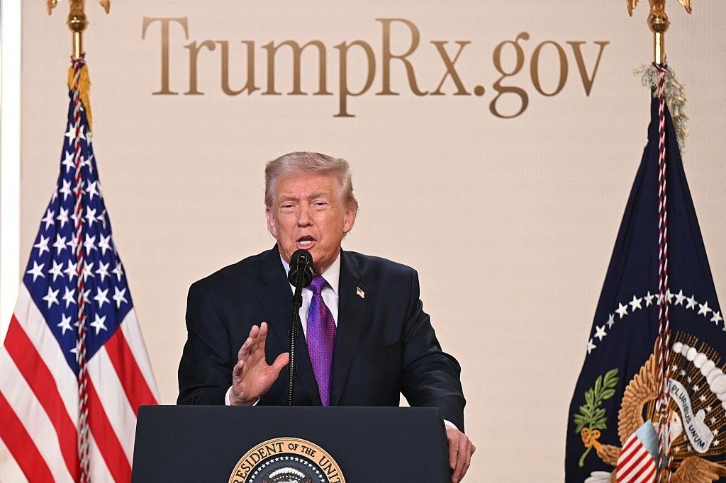 Person som taler ved et podium med to amerikanske flagg og et statlig insignier, foran et bakteppe med teksten
