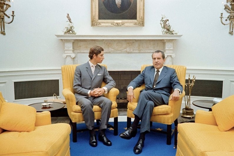 Two men successful  suits beryllium   connected  yellowish  chairs successful  an authoritative  room, engaged successful  conversation. A coating  and a fireplace are successful  the background