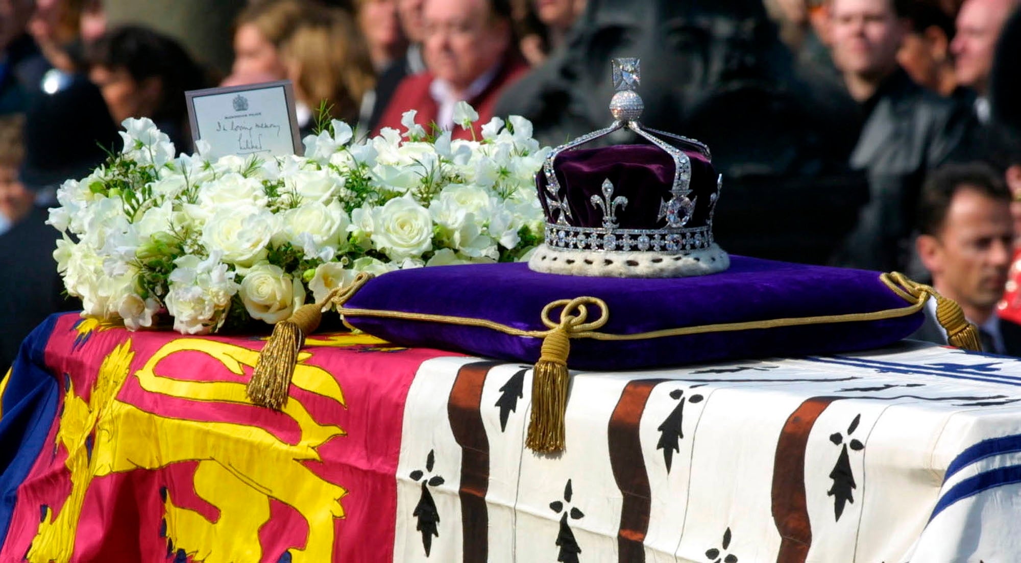 Close-up of a royal casket draped successful  flags, adorned with a crown, orb, and scepter, surrounded by achromatic  flowers