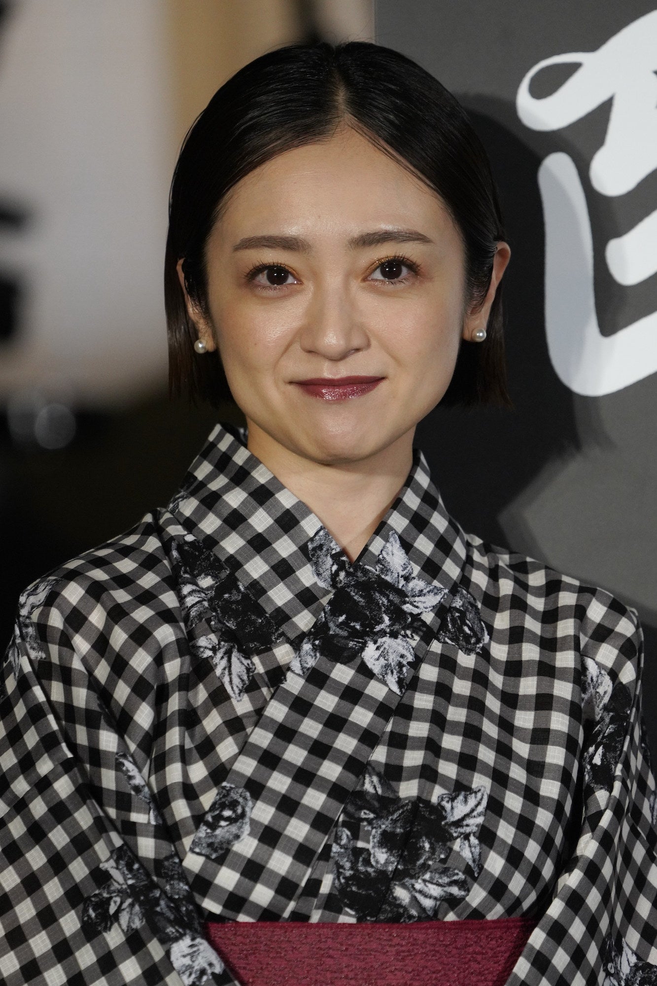 女性がチェック柄の着物と花柄の帯を身に着け、微笑んでいる。背景には漢字が見える。