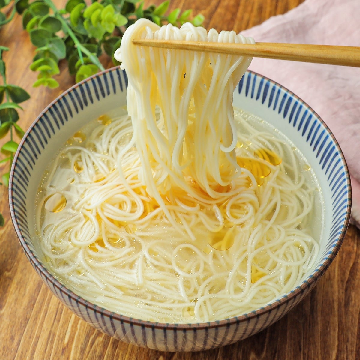 箸で白い麺を持ち上げている様子。丼にはスープと麺が入っている。背景に緑の葉が見える。