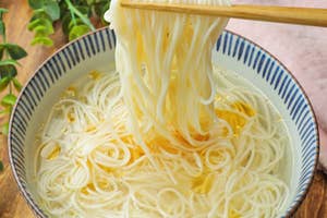 箸で白い麺を持ち上げている様子。丼にはスープと麺が入っている。背景に緑の葉が見える。