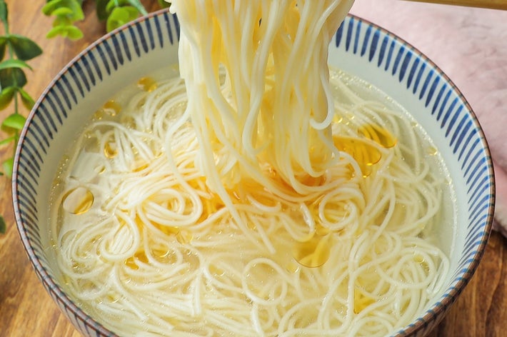 箸で白い麺を持ち上げている様子。丼にはスープと麺が入っている。背景に緑の葉が見える。