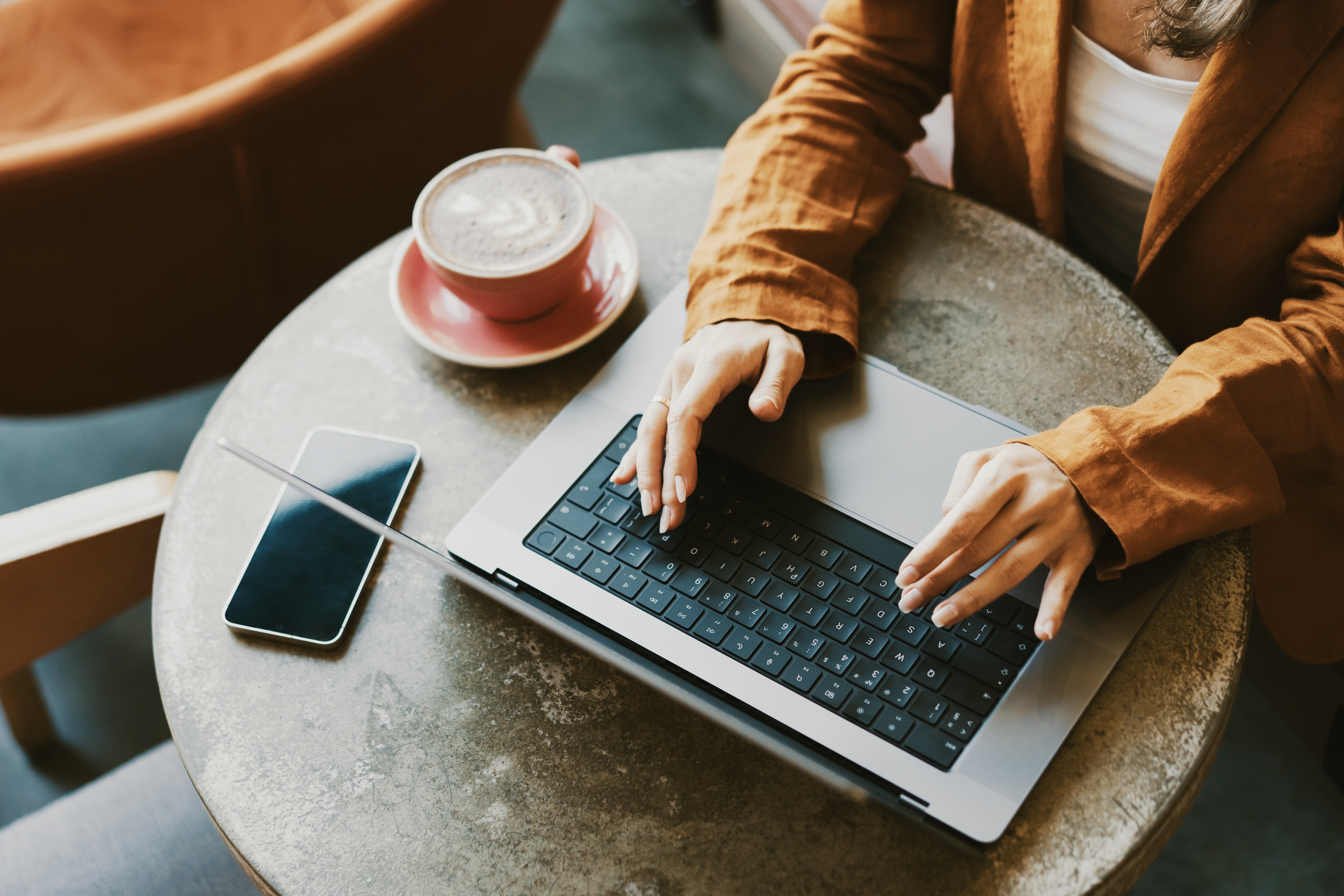 Person in a brown jacket typing on a laptop at a round table with a smartphone and coffee cup nearby