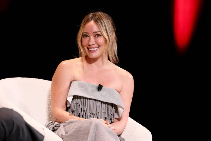 A pistillate   successful  a strapless pleated formal  sits connected  a achromatic  chair, smiling and speaking into a lapel microphone