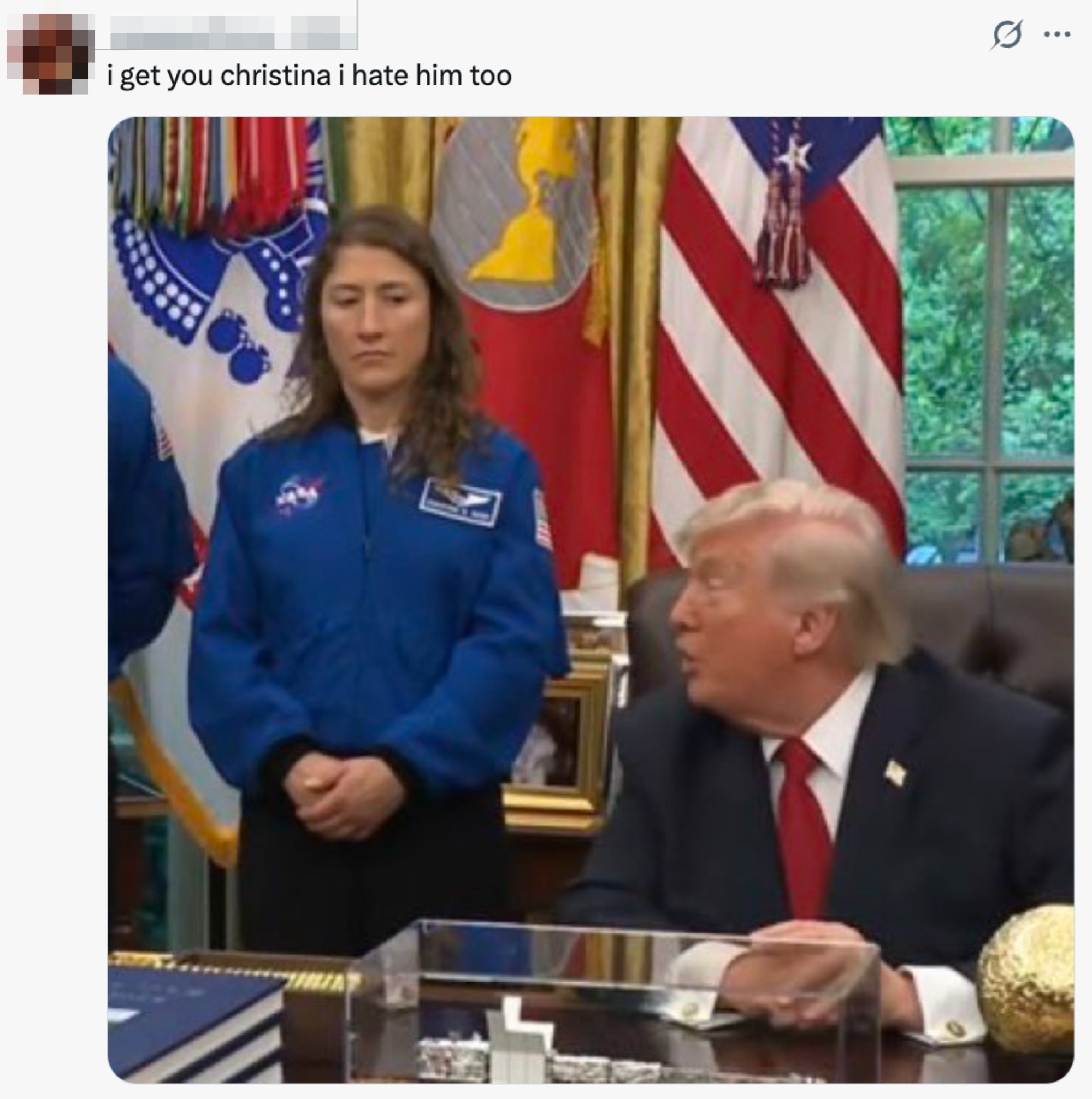 En astronaut i blå NASA-drakt står ved siden av en sittende mann i dress inne i et offisielt rom med amerikanske flagg