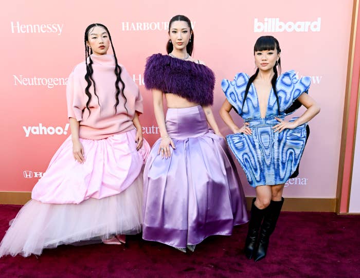 Three individuals airs  connected  a reddish  carpet. Each wears unique, avant-garde outfits with melodramatic  silhouettes and textures