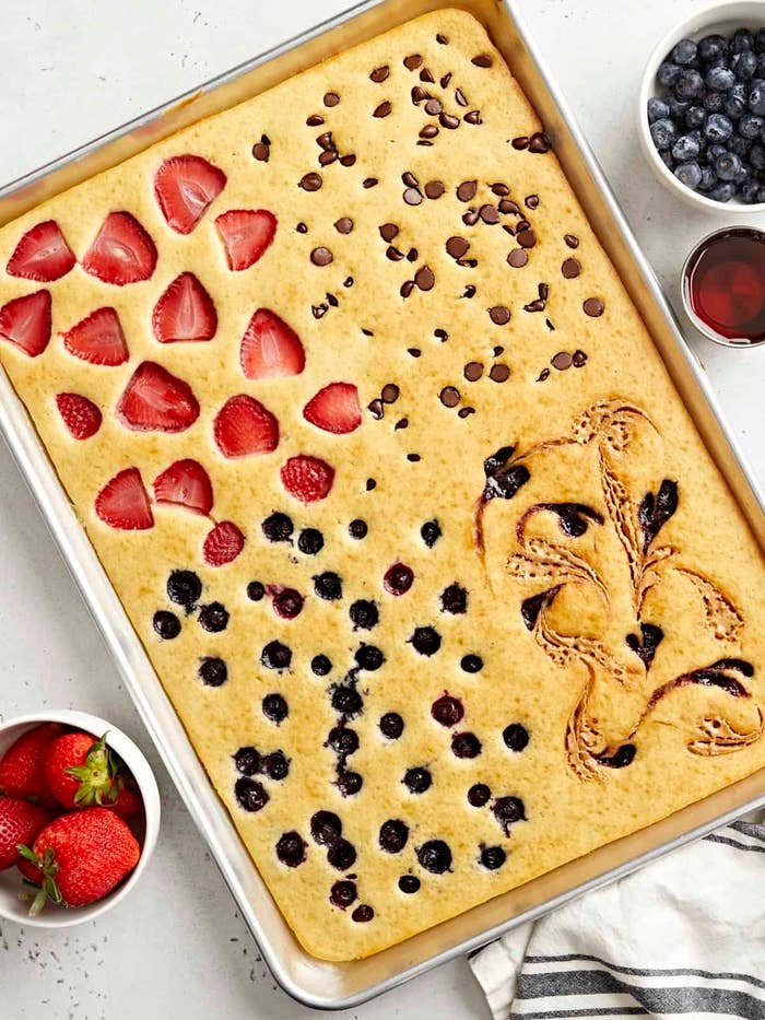 Large sheet pancake with sections of strawberries, chocolate chips, blueberries, and a decorative design. Bowls of blueberries and syrup nearby
