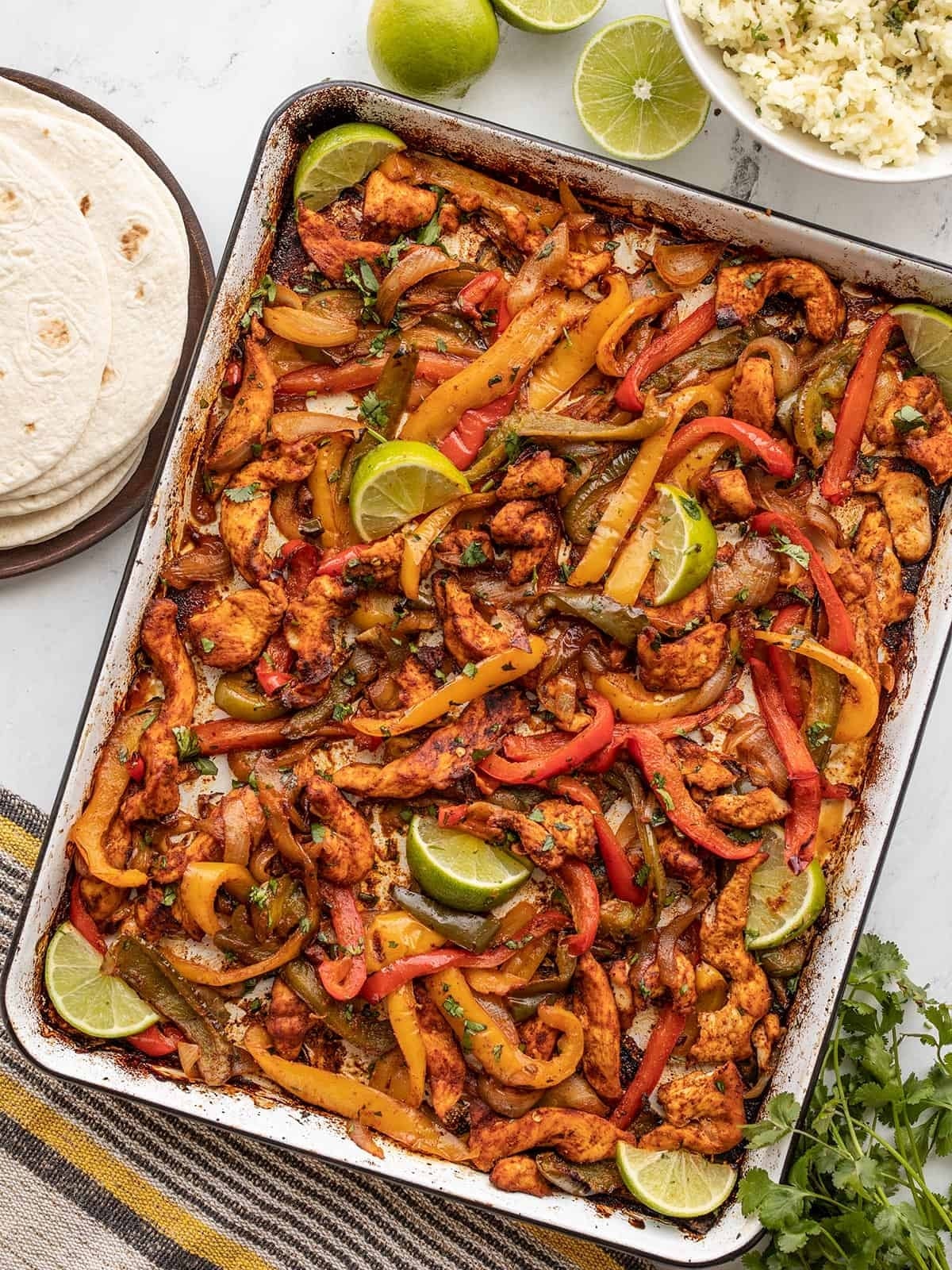 A tray of chicken fajitas with sliced bell peppers and lime wedges, tortillas, and a bowl of rice in the background