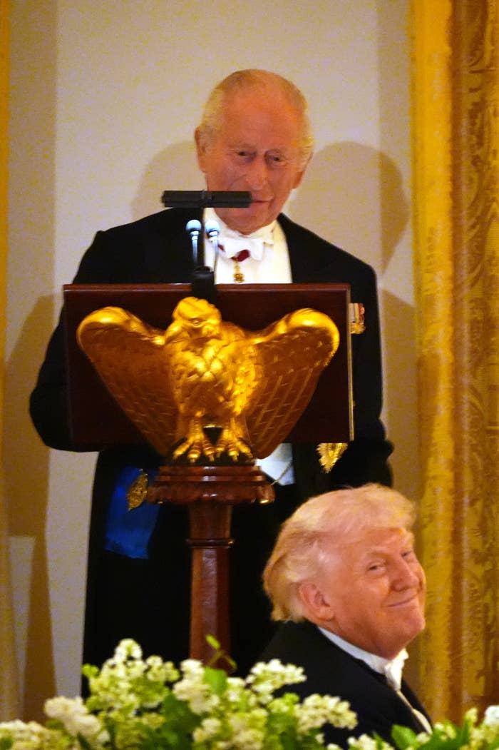 A antheral   speaks astatine  a podium with an eagle emblem, wearing ceremonial  attire. Another man, seated and smiling, glances to the side
