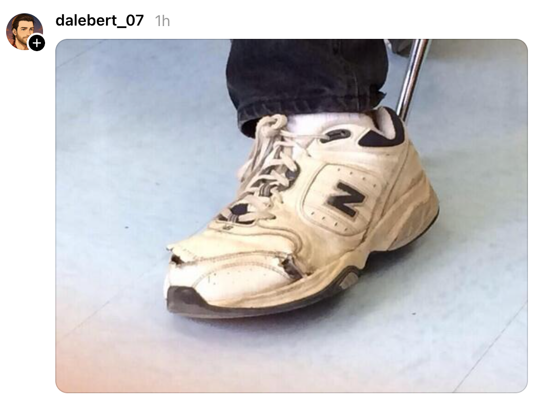 Sepatu kets putih usang dengan bagian depan robek dan terlihat usang, dikenakan oleh orang yang duduk di kursi