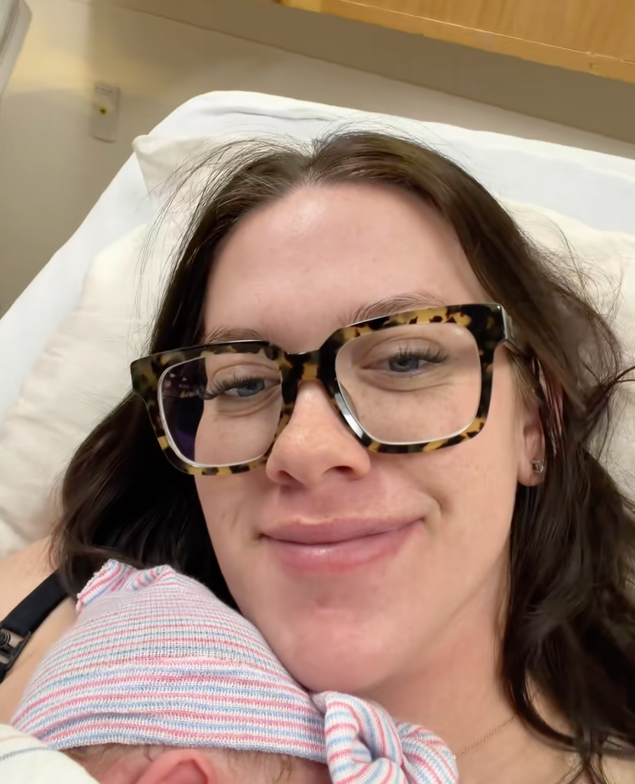 Person wearing glasses smiles portion    mildly  cradling a newborn wrapped successful  a striped broad  against their shoulder