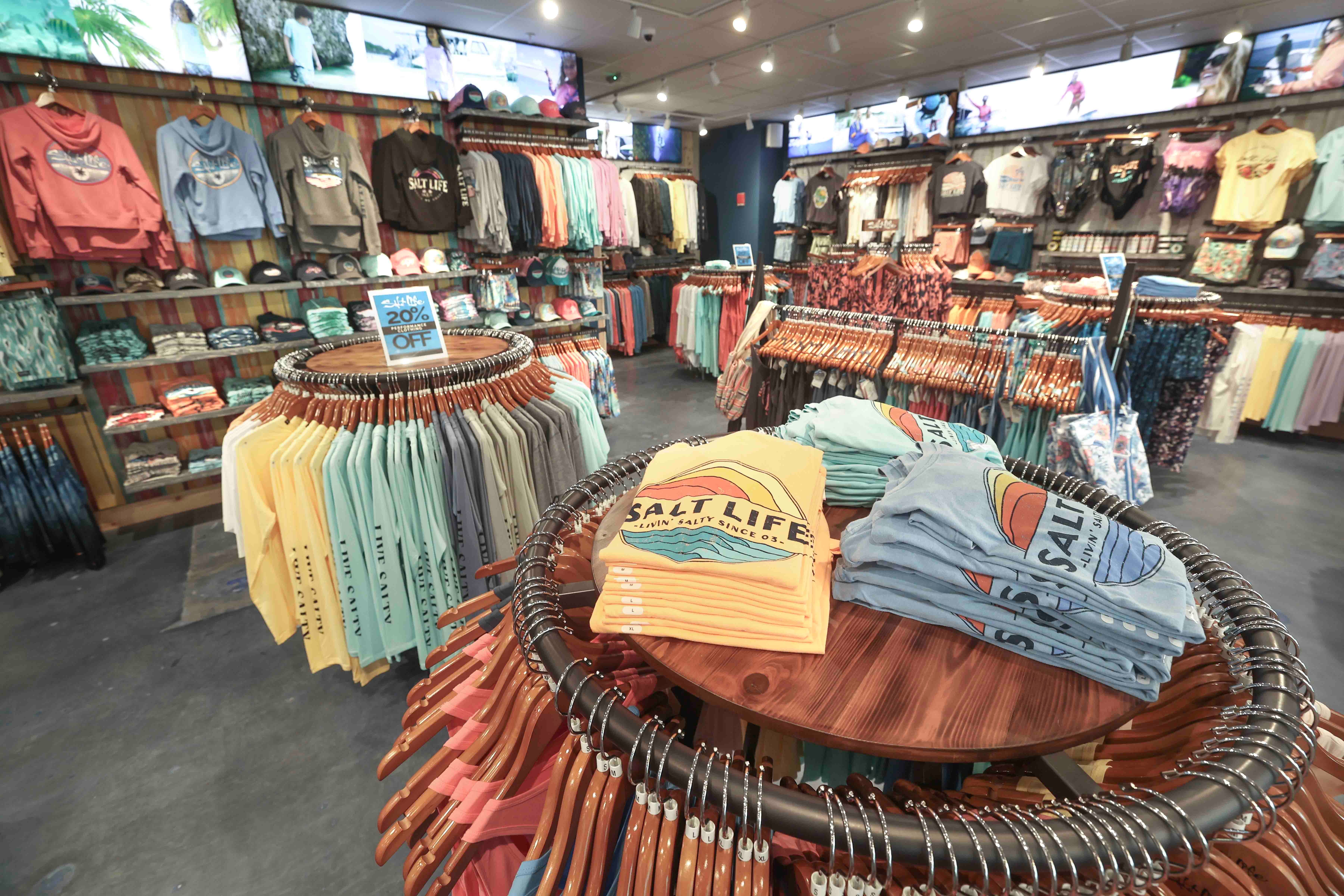 Interior toko pakaian dengan kaos grafis terlipat di meja pajangan, dikelilingi rak berbagai item pakaian. Tanda penjualan di atas meja