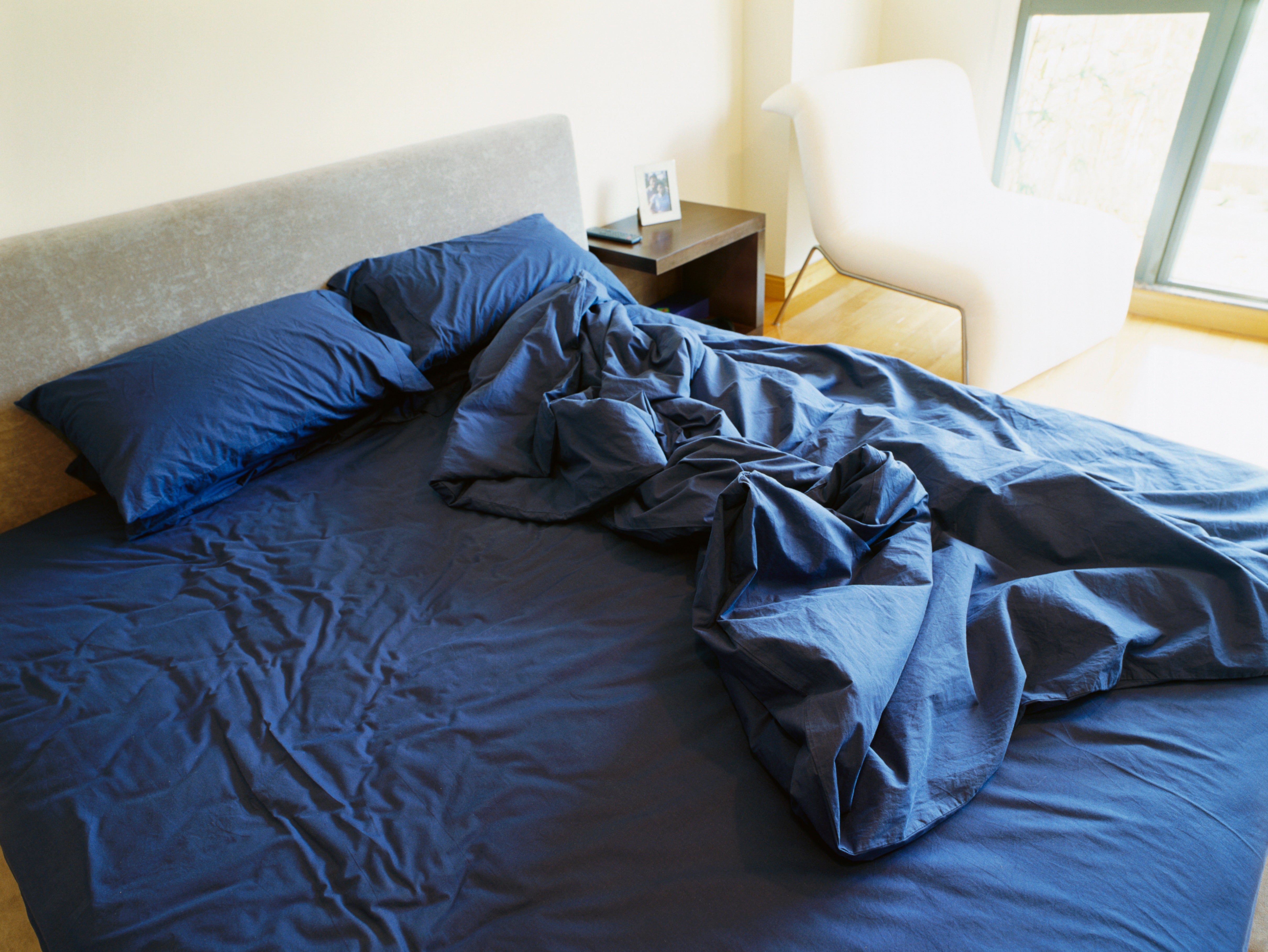 Tempat tidur yang belum dirapikan dengan seprai dan bantal kusut di ruangan yang diterangi matahari, di samping meja samping tempat tidur dengan foto berbingkai