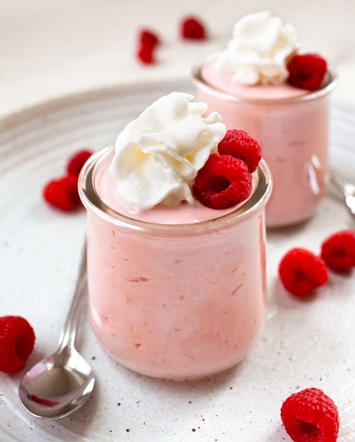 Two small jars of creamy pink mousse topped with whipped cream and raspberries, with spoons placed beside them