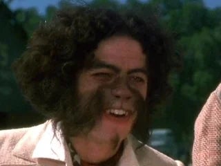 Person with curly hairsbreadth  and excessive facial hair, wearing a textured overgarment   and a collared shirt, outdoors