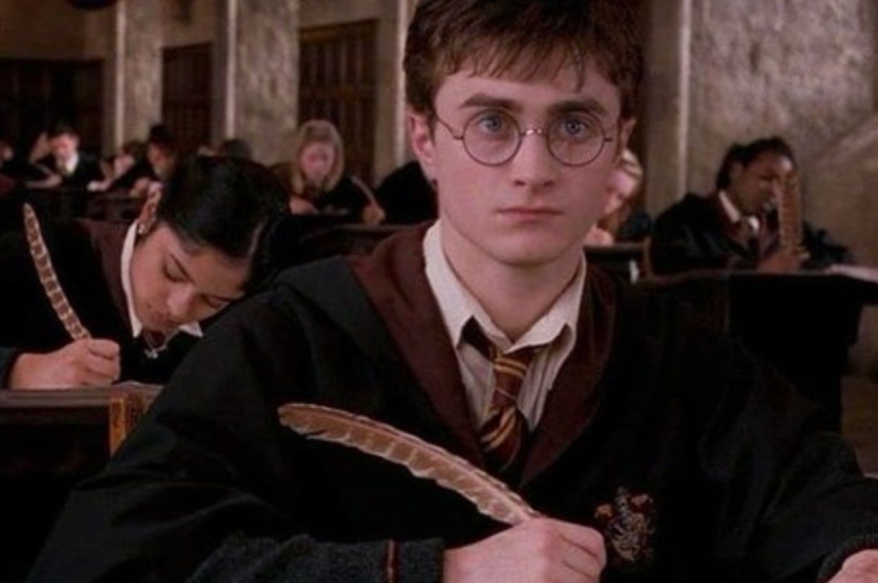 A young man with glasses in a school uniform sits in a classroom holding a quill pen, while a classmate writes behind him