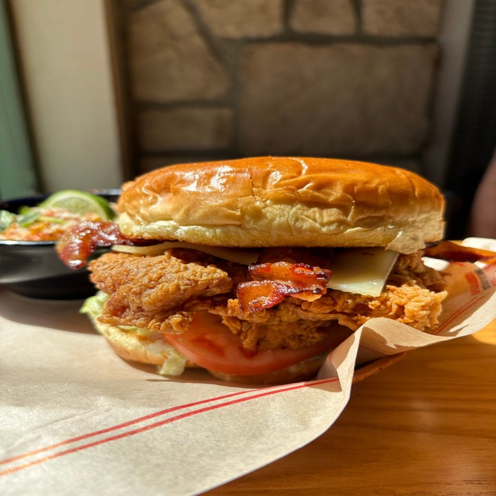 A crispy chicken sandwich with bacon, cheese, lettuce, and tomato on a bun, served on parchment paper with a side dish partly visible