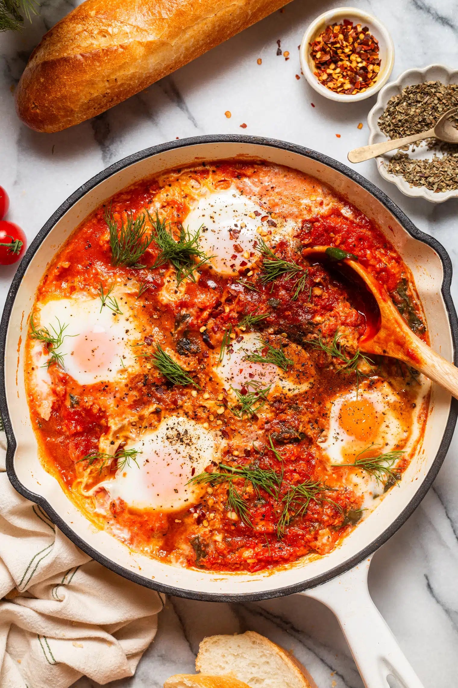 Skillet of shakshuka with poached eggs in tomato sauce, garnished with herbs, next to bread and spices on a marble surface