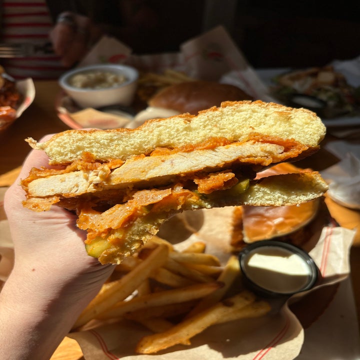 A hand holds a halved fried chicken sandwich with pickles, set above fries and sauce on a table