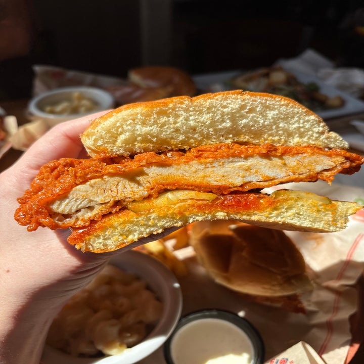 A hand holds a spicy fried chicken sandwich, cut in half, revealing the crispy chicken inside. Macaroni and cheese is visible in the background
