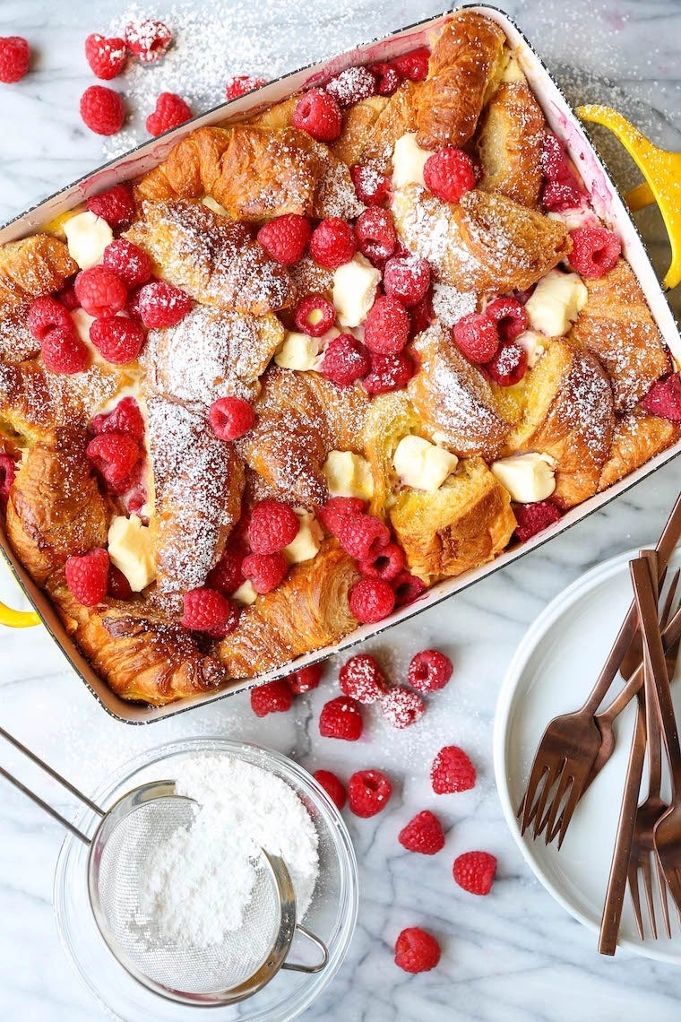 Baked croissant and raspberry dessert topped with powdered sugar, placed on a marble surface with additional raspberries beside a bowl and utensils