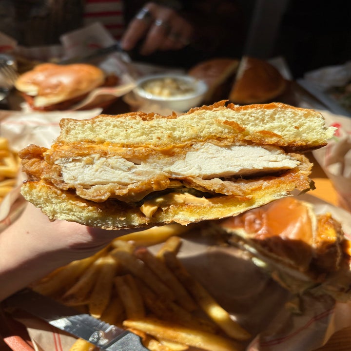 Close-up of a hand holding a crispy chicken sandwich with visible layers of fried chicken and sauce, with fries and another sandwich in the background