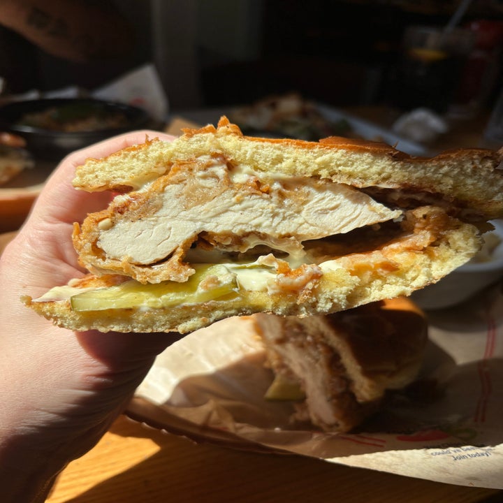 Close-up of a hand holding a fried chicken sandwich with pickles and a bite taken out, revealing the crispy chicken inside