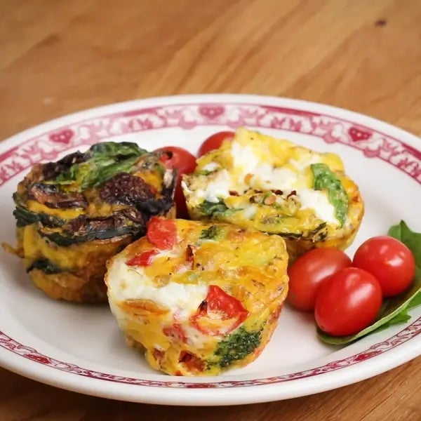 Three mini vegetable frittatas on a plate with cherry tomatoes and spinach, arranged on a wooden table