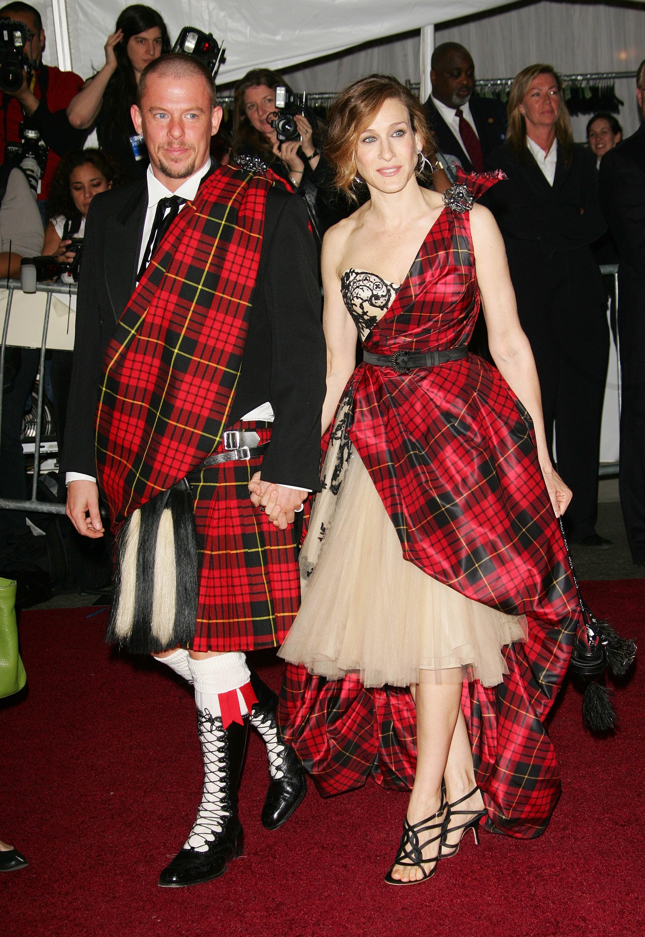 Two radical   connected  a reddish  carpet, wearing matching tartan plaid outfits. The woman’s formal  has an asymmetrical plan  with a tulle skirt
