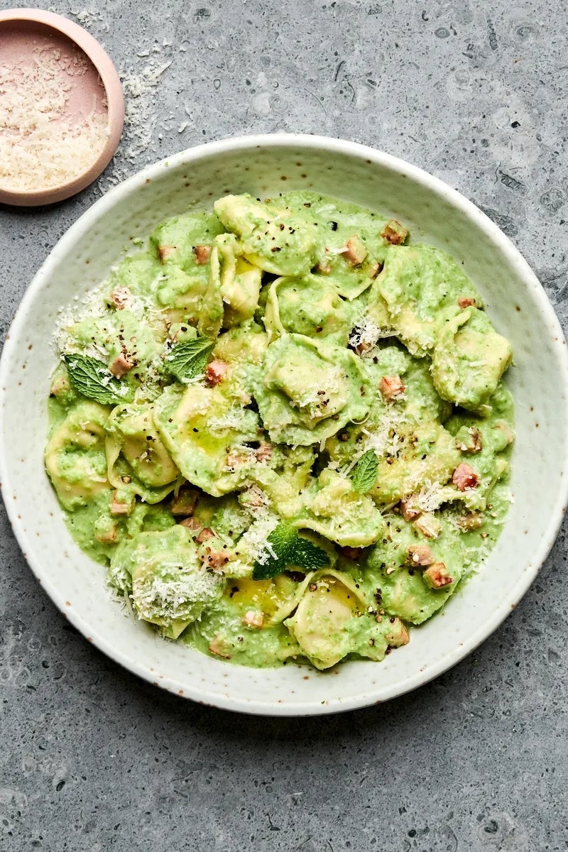A dish of creamy green pasta garnished with cheese, mint leaves, and diced pieces on a speckled plate