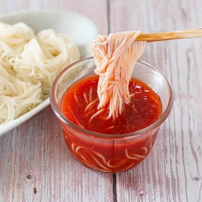 ガラス容器に入った赤いソースに麺を浸す様子。隣に茹でられた麺が見える。