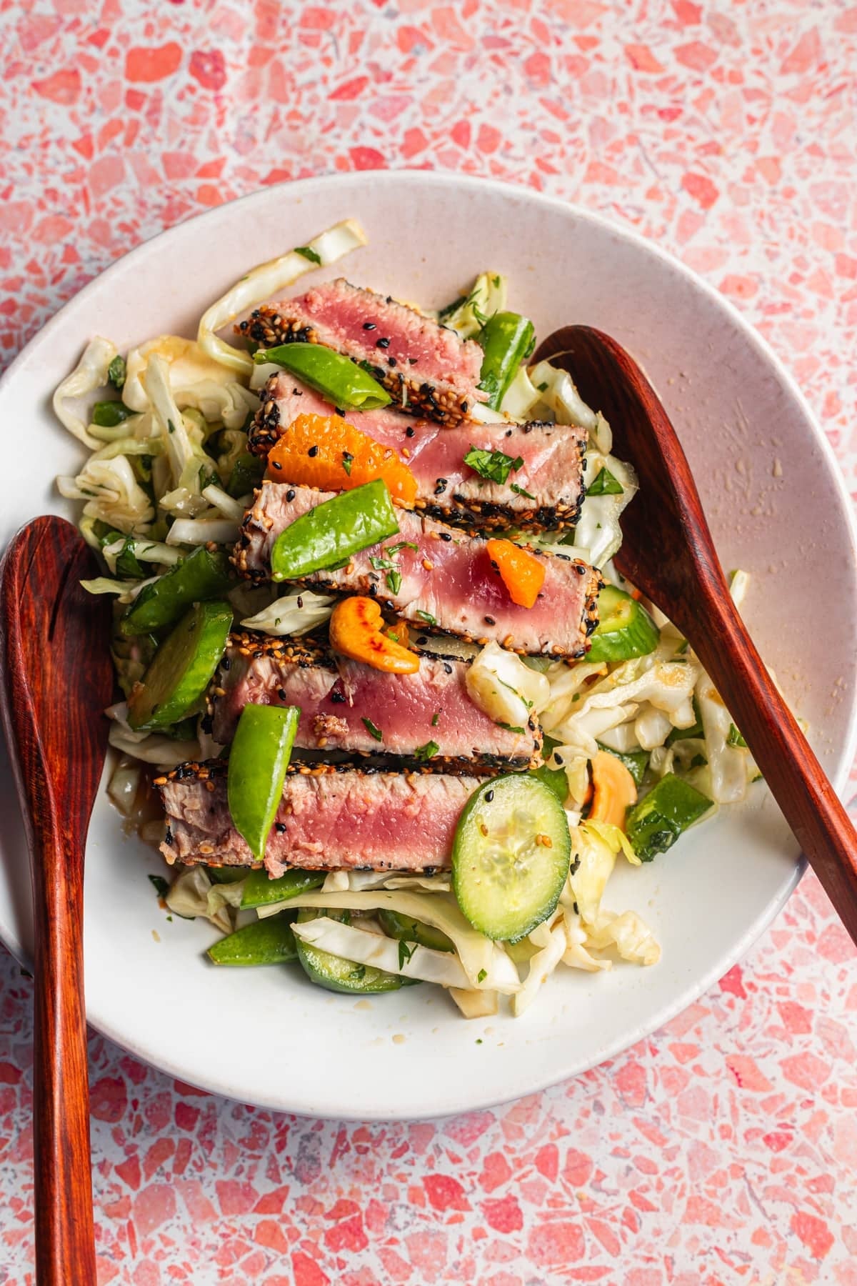 Seared tuna slices on a bed of mixed cabbage and vegetables, served with wooden salad utensils in a white bowl