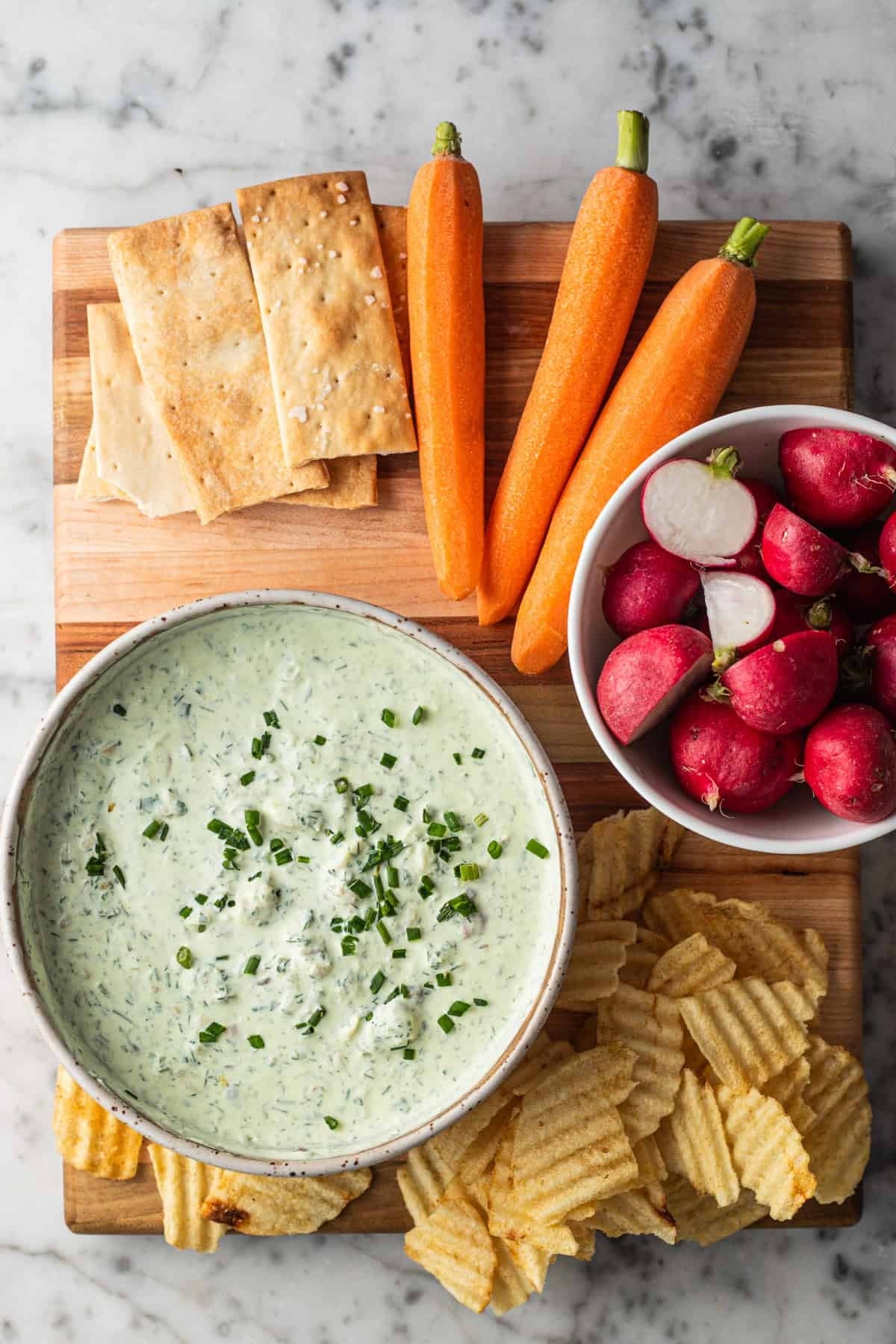 A wooden board with a bowl of green herb dip, surrounded by crackers, carrots, radishes, and ridged potato chips
