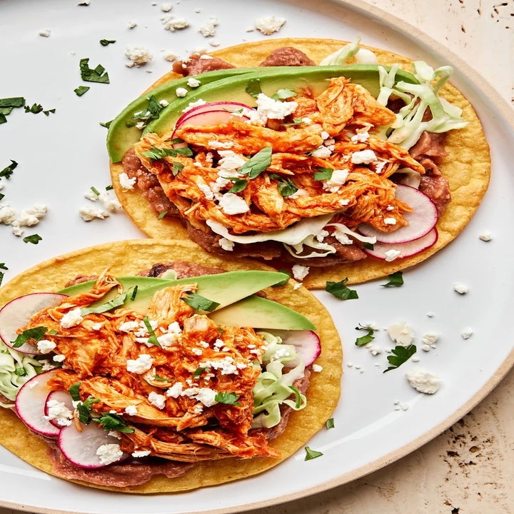 Two tostadas topped with shredded chicken, sliced avocado, radishes, cabbage, and crumbled cheese on a white plate
