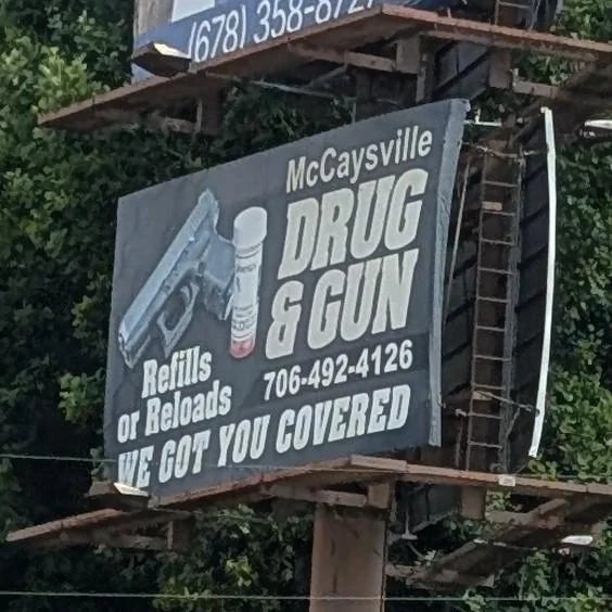 Billboard for McCaysville Drug &amp;amp; Gun, showing a weapon  and pill bottle. It states "Refills oregon  Reloads" and "We Got You Covered," with a telephone  number