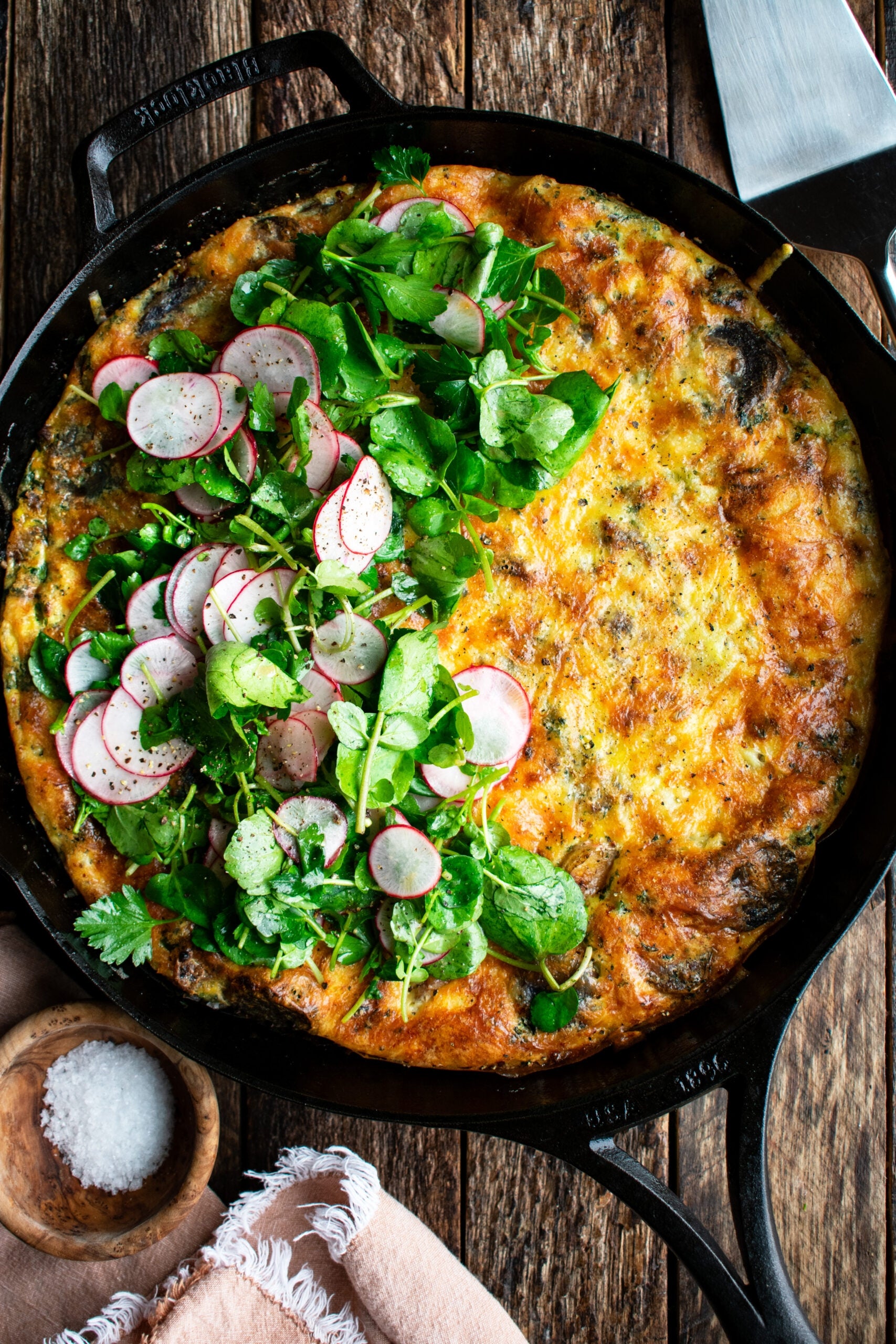 Frittata in a skillet topped with sliced radishes and fresh greens, on a wooden table with a small bowl of salt